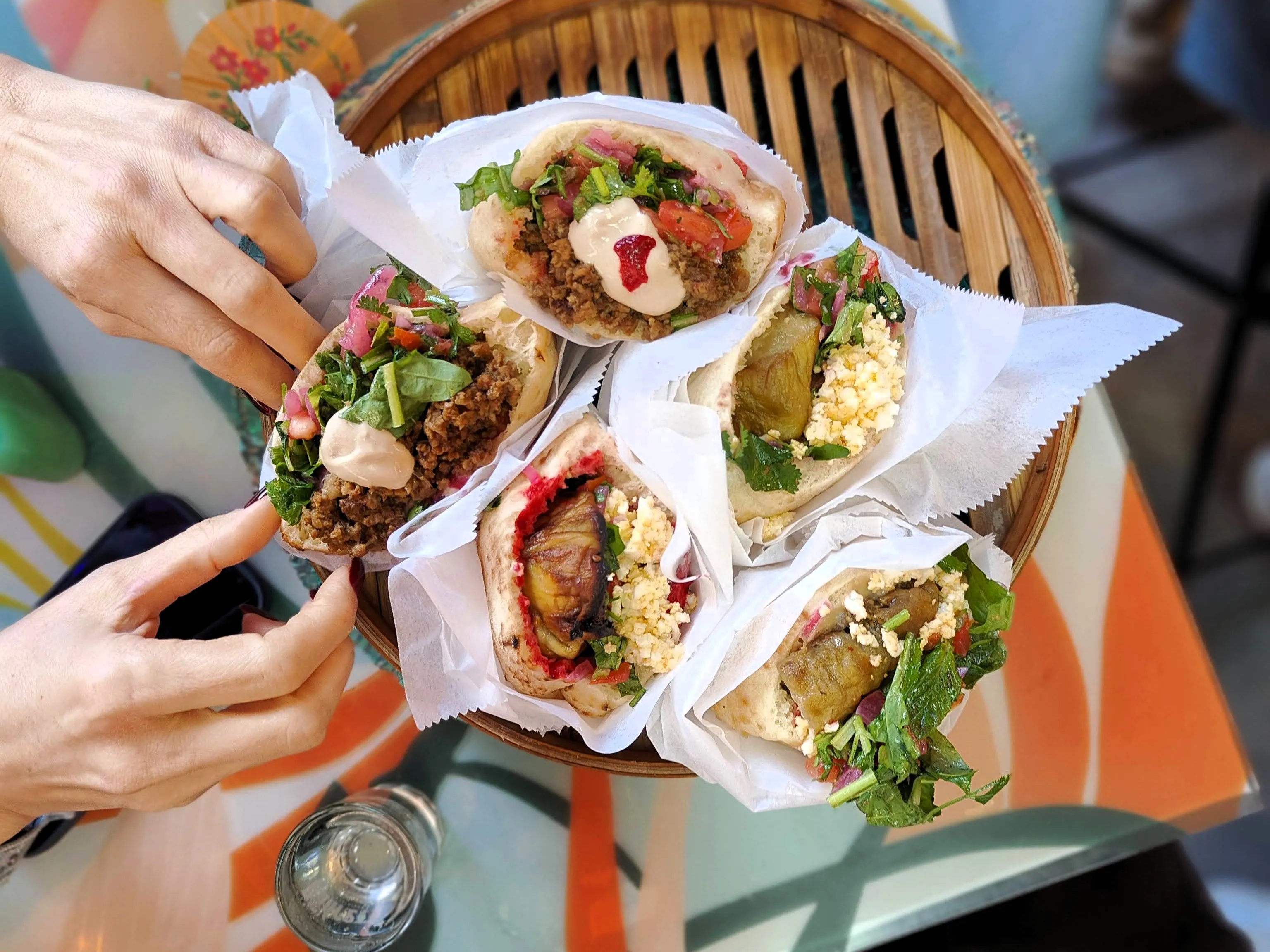 Two people reaching for a basket of overflowing pita sandwiches with fresh herbs, tahini, and roasted vegetables at Carmel Market, Tel Aviv