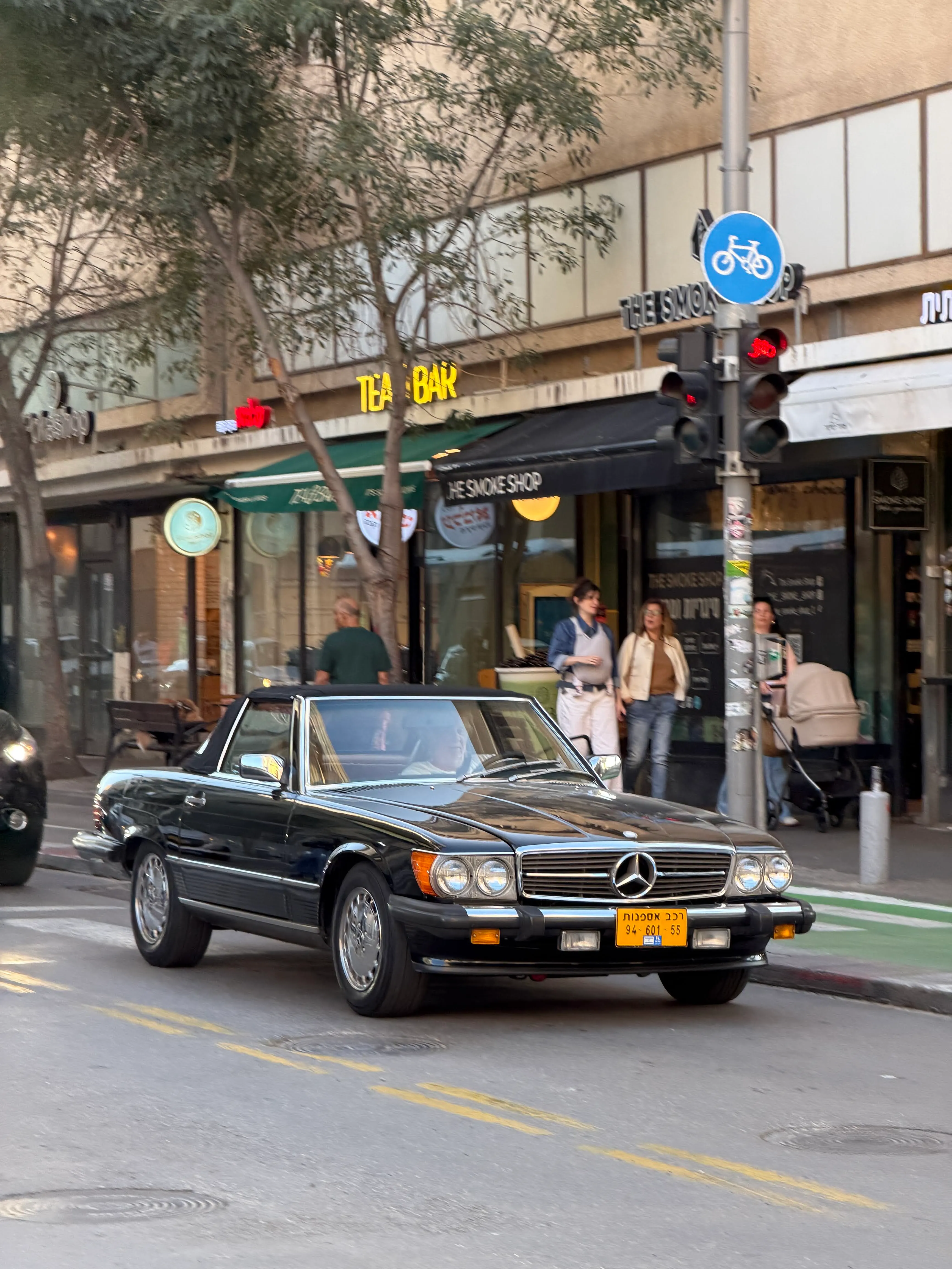 A classic vintage Mercedes convertible on a tree-lined Tel Aviv street, with cafés, shops, and pedestrians in the late afternoon light